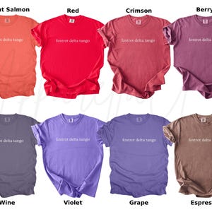 Op de afbeelding: Acht verschillende gekleurde t-shirts worden getoond, waaronder helder zalm, rood, karmozijnrood, bes, wijn, violet, druif en espresso. Elk shirt heeft de tekst "foxtrot delta tango" op de voorkant.