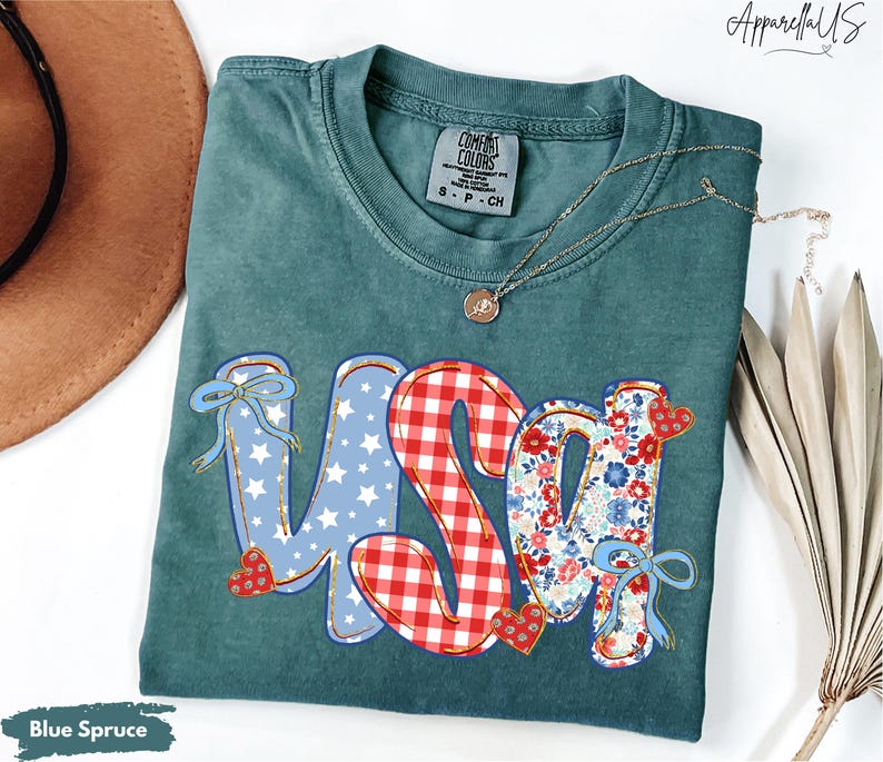 May include: Blue Spruce Comfort Colors t-shirt with a USA graphic. The letters are decorated with patriotic patterns: blue stars, red gingham, and floral prints. A gold necklace with a pendant rests on the shirt.