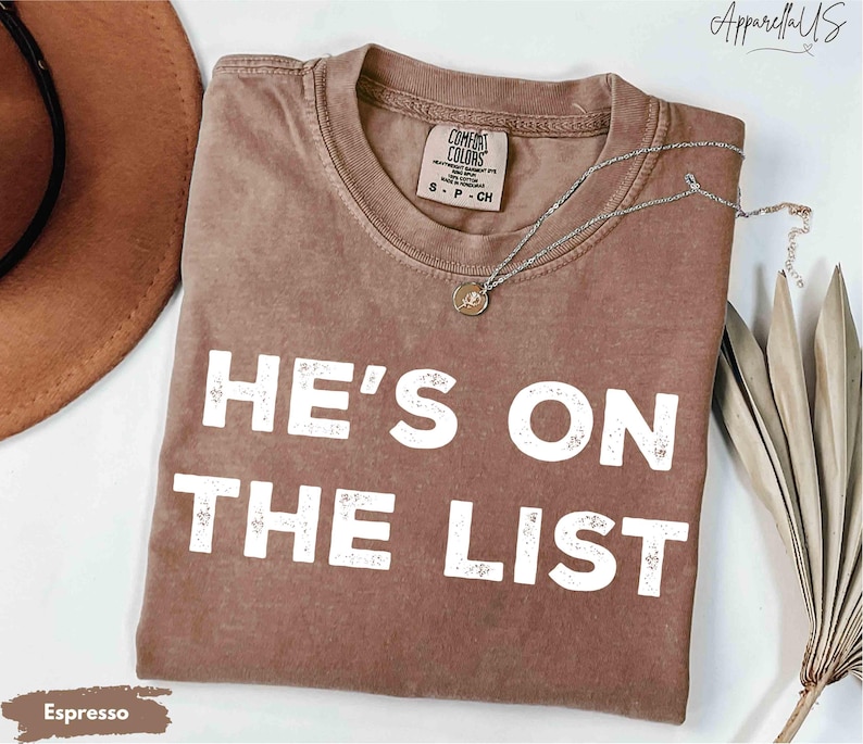 May include: Espresso-coloured t-shirt with the text "HE'S ON THE LIST" in white block letters. The shirt has a crew neck and short sleeves. A silver necklace with a small pendant is draped across the shirt. The shirt is folded on a white surface.