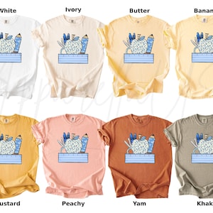 May include: Ten different colored t-shirts with a graphic of a blue apple with pencils and a ruler. The colors are white, ivory, butter, banana, mustard, peachy, yam, khaki, and black.