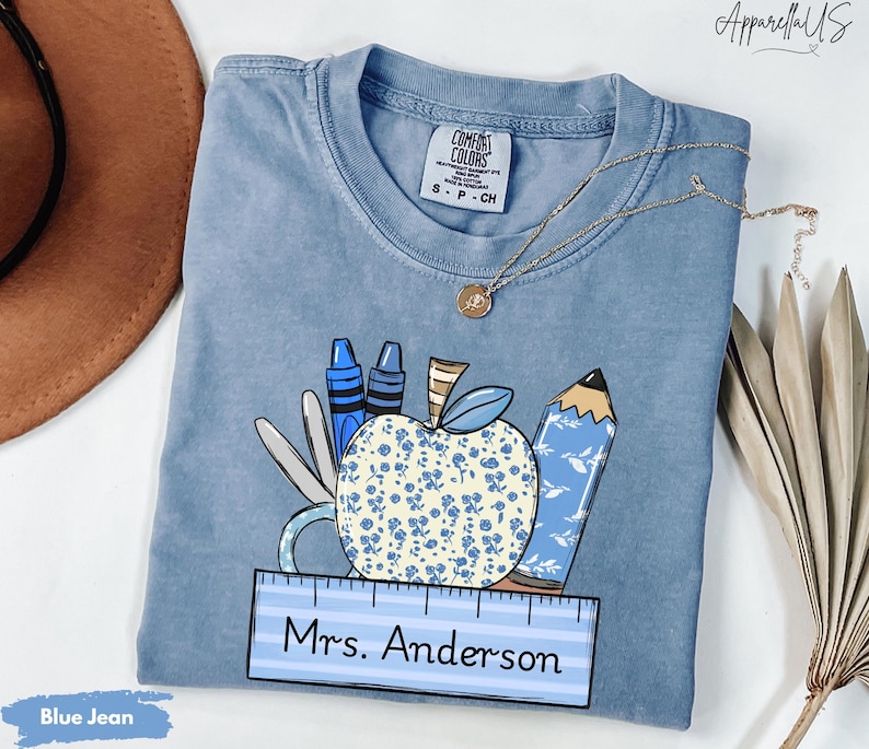 May include: Blue jean t-shirt with a graphic of a blue apple with white flowers, crayons, a pencil, and a ruler. The text "Mrs. Anderson" is on the ruler.