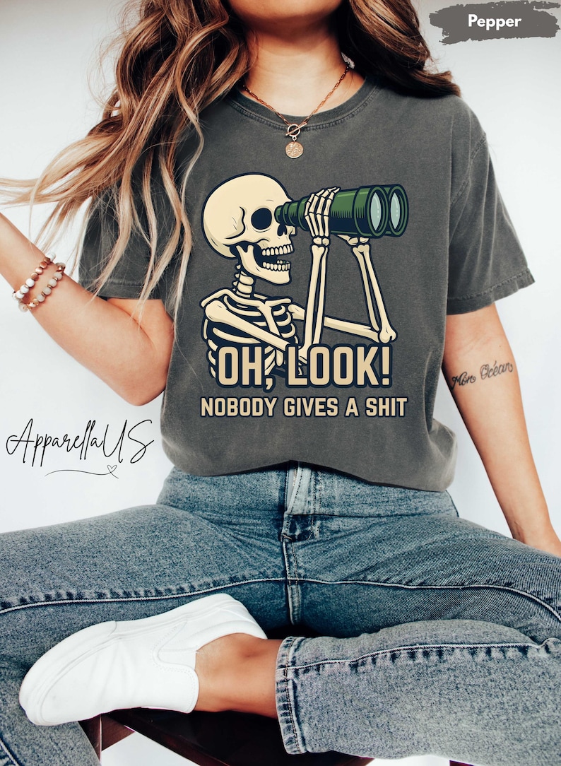 Op de afbeelding: Donkergrijs T-shirt met een skelet dat door een verrekijker kijkt. Het skelet houdt de verrekijker met beide handen vast. De tekst op het shirt luidt "OH LOOK! NOBODY GIVES A SHIT".