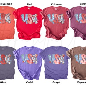 May include: A collection of eight t-shirts in various colors, each featuring the word "USA" in a patriotic design. The letters are a mix of blue polka dots and red gingham patterns. Colors include bright salmon, red, crimson, berry, wine, violet, grape, and espresso.