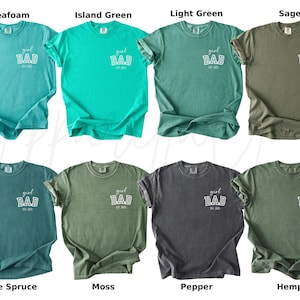 May include: A collection of eight t-shirts in various colors, including seafoam, island green, light green, sage, blue spruce, moss, pepper, and hemp. Each shirt has the text "girl DAD est. 2023" printed on the front.