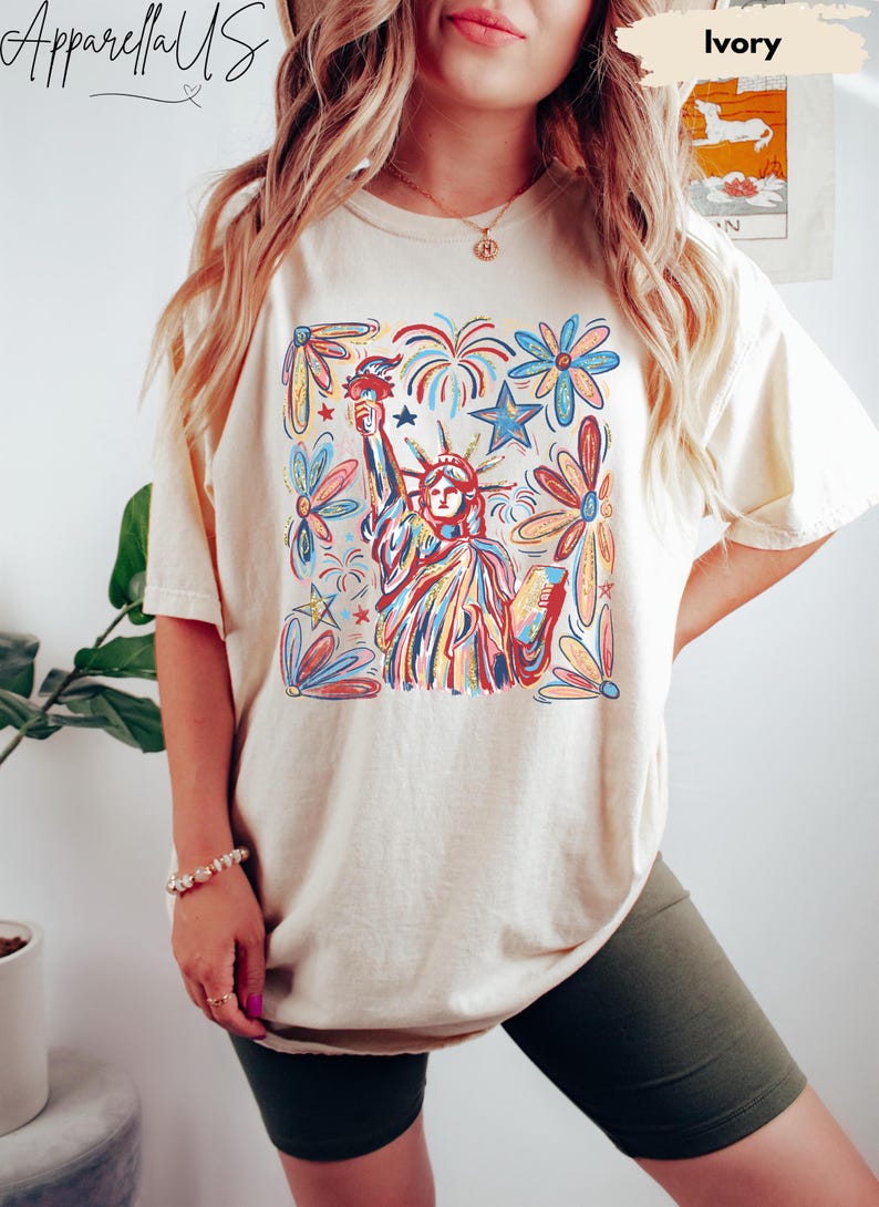 Puede incluir: Camiseta de color marfil con una colorida ilustraci&oacute;n de la Estatua de la Libertad con fuegos artificiales y elementos florales. La camiseta tiene mangas cortas y un corte holgado. El texto "Apparell US" es visible en la parte superior izquierda.