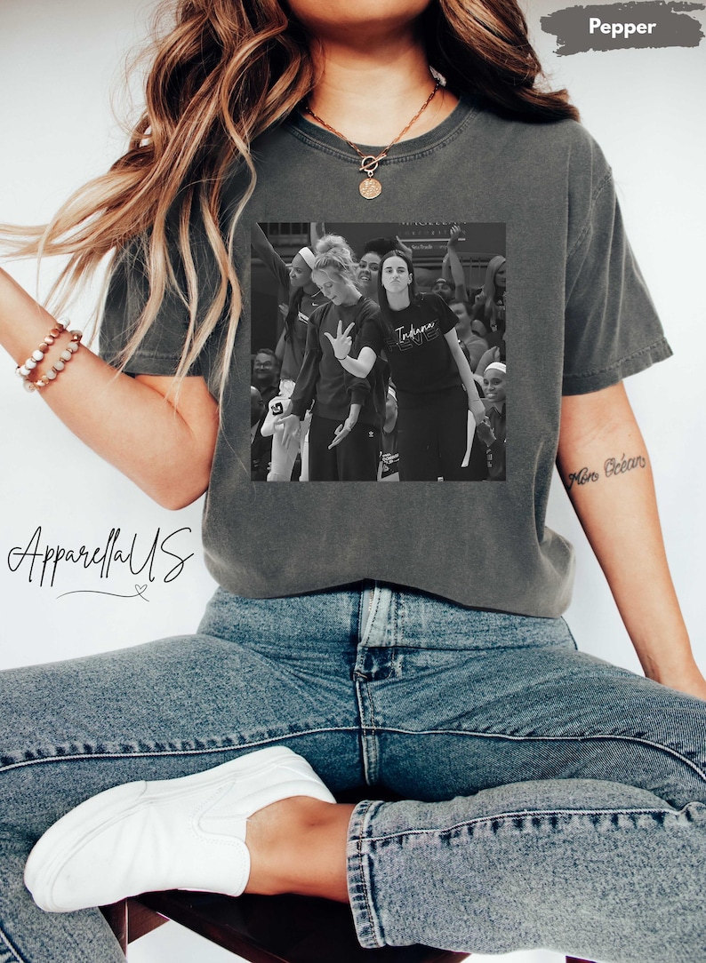 May include: A gray t-shirt with a black and white image of a basketball team. The shirt has the word "Indiana" printed on it. The person is wearing blue jeans and white shoes. The text "ApparellaUS" is on the image.
