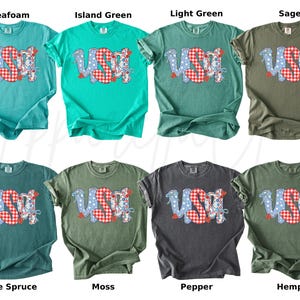 May include: A collection of eight t-shirts in various colors, each featuring the word "USA" in a patriotic design. The letters are a mix of blue polka dots and red gingham patterns. The shirts are in shades of green, blue, and gray.