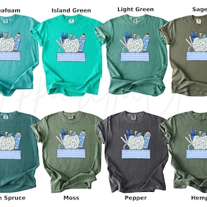 May include: A set of ten t-shirts featuring a graphic of a red apple with school supplies, including pencils, scissors, and a ruler. The shirts are available in ten different colors: Seafoam, Island Green, Light Green, Sage, Blue Spruce, Moss, Pepper, Hemp, and two more.