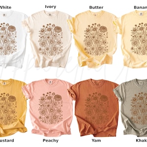 Pode incluir: Uma cole&ccedil;&atilde;o de oito camisetas de manga curta em v&aacute;rias cores: branco, marfim, manteiga, banana, mostarda, p&ecirc;ssego, inhame e c&aacute;qui. Cada camisa apresenta um desenho floral marrom na frente, com os nomes das cores abaixo.