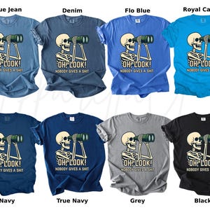 Op de afbeelding: Een collectie t-shirts in diverse kleuren, elk met een skelet dat door een verrekijker kijkt. De tekst "OH LOOK! NOBODY GIVES A SHIT" staat eronder. Kleuren zijn blue jean, denim, flo blue, royal caribe, navy, true navy, grijs en zwart.
