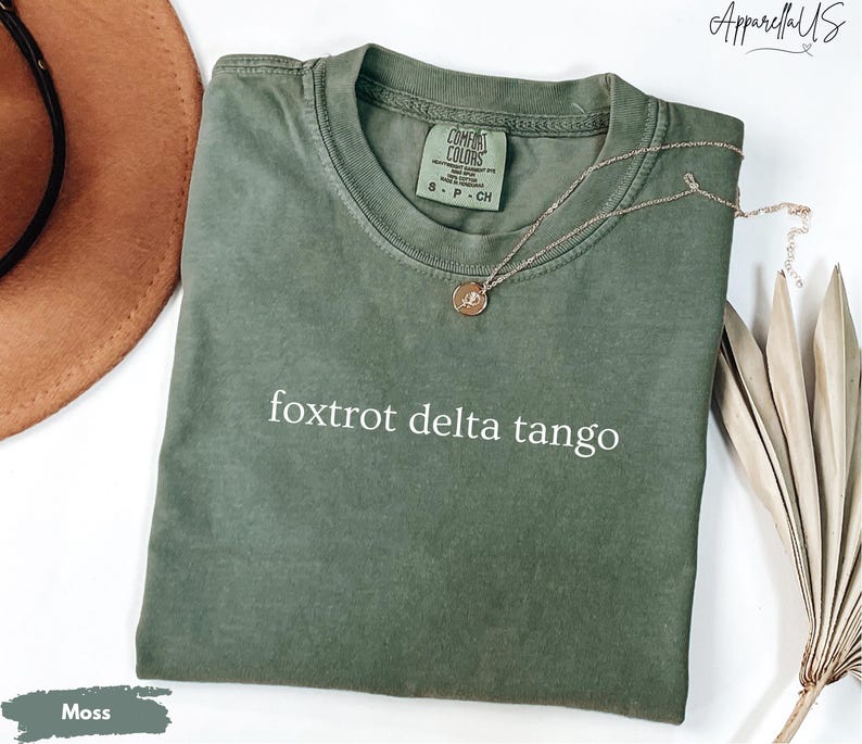 Op de afbeelding: Een mosgroen t-shirt met de tekst "foxtrot delta tango" in het wit. Het shirt heeft een ronde hals en korte mouwen. Een gouden ketting met een hanger is over het shirt gedrapeerd. Een bruine hoed en een gedroogde plant staan op de achtergrond.