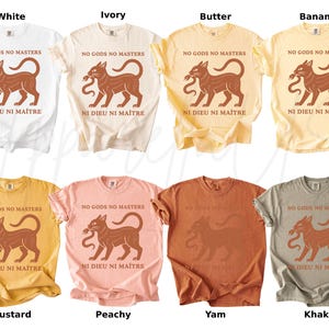 May include: A collection of eight t-shirts in various colors, including white, ivory, butter, banana, mustard, peachy, yam, and khaki. Each shirt features a brown graphic of a cat holding a snake, with the text "NO GODS NO MASTERS" and "NI DIEU NI MA&Icirc;TRE".