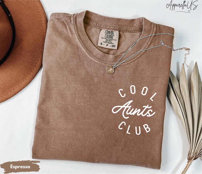 May include: A brown t-shirt with the text "COOL AUNTS CLUB" in white lettering. The shirt is made of a soft, comfortable material and has a relaxed fit.