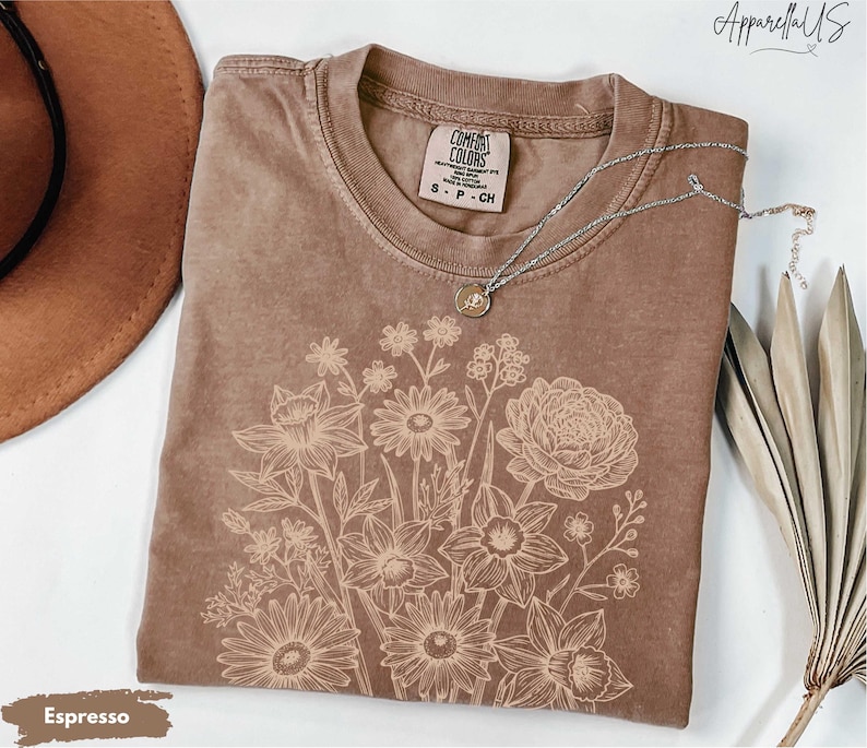 Pode incluir: Camiseta cor de caf&eacute; com um design floral. A camiseta tem gola redonda e mangas curtas. Um colar de prata com um pingente est&aacute; drapeado sobre a camiseta. A camiseta est&aacute; rotulada como "Comfort Colors".