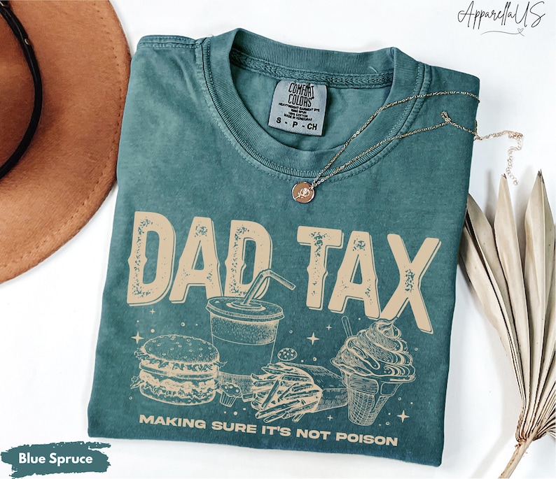 May include: Blue spruce t-shirt with the words "DAD TAX" in a distressed font, along with illustrations of a burger, fries, soda, and ice cream cone. The text "MAKING SURE IT'S NOT POISON" is below the graphic. A gold necklace is draped on the shirt.