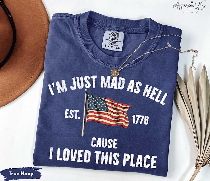 May include: True navy blue t-shirt with the text "I'M JUST MAD AS HELL CAUSE I LOVED THIS PLACE" and an American flag graphic. The shirt includes the year "1776" and the brand "Comfort Colors". A gold necklace is draped across the shirt.