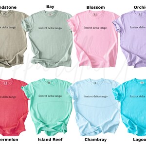 Op de afbeelding: Acht T-shirts met korte mouwen in verschillende kleuren, waaronder beige, lichtgroen, roze en paars. Elk shirt heeft de tekst "foxtrot delta tango" op de borst gedrukt. Andere kleuren zijn rood, teal, lichtblauw en turquoise.