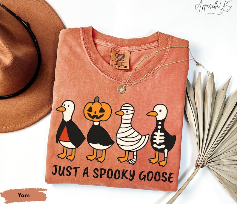 K&ouml;nnte beinhalten: Ein yam-farbenes T-Shirt mit Rundhalsausschnitt und einem Halloween-Motiv. Die Grafik zeigt vier Cartoon-G&auml;nse, die als Vampir, K&uuml;rbis, Mumie und Skelett verkleidet sind. Der Text "JUST A SPOOKY GOOSE" ist darunter gedruckt.