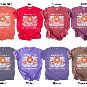 May include: A collection of eight t-shirts in various colors, including bright salmon, red, crimson, berry, wine, violet, grape, and espresso. Each shirt features the text "ABOLISH ICE" and "NO HUMAN IS ILLEGAL" with a floral design.