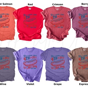 May include: Assorted-color t-shirts with a vintage design for the 4th of July. Each shirt features a hot dog graphic and the text "You look like 4th of July makes me a real bad hot dog". Colors include bright salmon, red, crimson, berry, wine, violet, grape, and espresso.