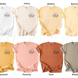 May include: A collection of eight short-sleeved t-shirts in various colors, including white, ivory, butter, banana, mustard, peachy, yam, and khaki. Each shirt features the text "girl DAD est. 2023" printed on the front.