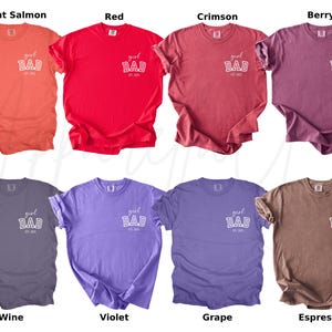May include: A collection of eight solid-color t-shirts in various shades, including bright salmon, red, crimson, berry, wine, violet, grape, and espresso. Each shirt features the text "girl DAD est. 2023" printed on the front.