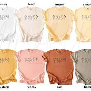May include: Ten different colour t-shirts with a simple floral design printed on the front. The colours are white, ivory, butter, banana, mustard, peachy, yam, khaki, and two shades of grey.