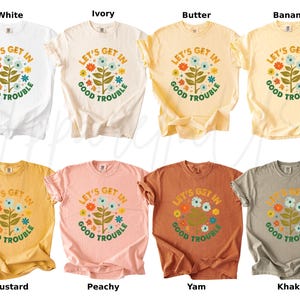 Peut inclure: Huit t-shirts de diff&eacute;rentes couleurs, dont blanc, ivoire, beurre, banane, moutarde, p&ecirc;che, igname et kaki. Chaque t-shirt pr&eacute;sente un motif floral avec le texte "LET'S GET IN GOOD TROUBLE" dans un style r&eacute;tro.