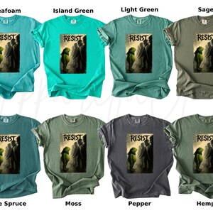 Pu&ograve; includere: Una collezione di t-shirt in vari colori, tra cui seafoam, verde isola, verde chiaro, salvia, abete blu, muschio, pepe e canapa. Ogni maglietta presenta la parola "RESIST" sopra una grafica.