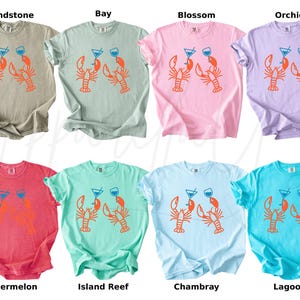 K&ouml;nnte beinhalten: Acht T-Shirts in verschiedenen Farben, jedes mit einem Siebdruckdesign von zwei Hummern, die Cocktails halten. Die Shirts sind in den Farbt&ouml;nen Beige, Gr&uuml;n, Rosa, Lila, Rot, T&uuml;rkis, Hellblau und T&uuml;rkis gehalten.