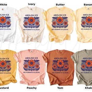 May include: A collection of t-shirts in various colors, including white, ivory, butter, banana, mustard, peachy, yam, and khaki. Each shirt features the text "ABOLISH ICE" above a floral design and the phrase "NO HUMAN IS ILLEGAL" below.