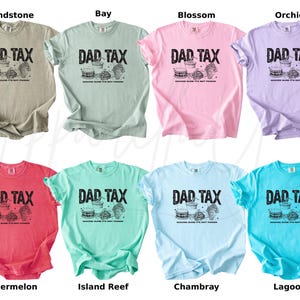 May include: A collection of eight short-sleeved t-shirts in various colors, including beige, green, pink, purple, red, teal, light blue, and turquoise. Each shirt features the text "DAD TAX" above a graphic of food items. The shirts are made of a soft material.