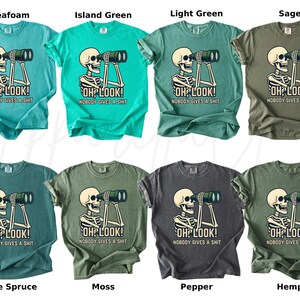 Op de afbeelding: Acht T-shirts in verschillende kleuren, elk met een skelet dat door een verrekijker kijkt. De tekst "OH LOOK! NOBODY GIVES A SHIT" staat eronder. Kleuren: seafoam, island green, light green, sage, blue spruce, moss, pepper en hemp. Afmetingen in cm.