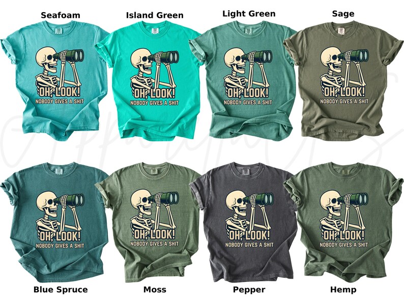 Op de afbeelding: Acht T-shirts in verschillende kleuren, elk met een skelet dat door een verrekijker kijkt. De tekst "OH LOOK! NOBODY GIVES A SHIT" staat eronder. Kleuren: seafoam, island green, light green, sage, blue spruce, moss, pepper en hemp. Afmetingen in cm.