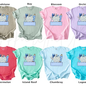 May include: Ten different color t-shirts with a graphic of a blue apple with school supplies around it and the text "First Day of School" printed on a blue banner below the apple.