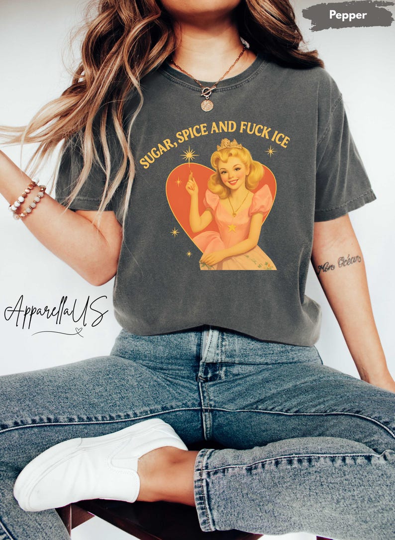 Peut inclure: T-shirt gris fonc&eacute; avec l'inscription dor&eacute;e "SUGAR, SPICE AND FUCK ICE". Le t-shirt pr&eacute;sente une illustration r&eacute;tro d'une femme en robe rose et couronne dans un c&oelig;ur. La marque "ApparellaUS" est visible.