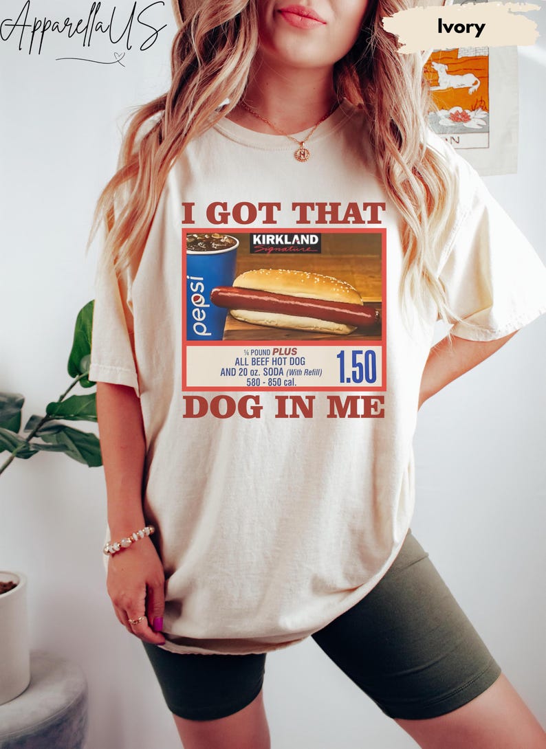 以下が含まれることがあります： 「I GOT THAT DOG IN ME」の文字が入ったアイボリー色のTシャツ。ホットドッグ、ペプシのカップ、カークランドのロゴが描かれています。Tシャツには、ホットドッグが&frac14;ポンドプラスのオールビーフホットドッグと20オンスのソーダであることも記載されています。