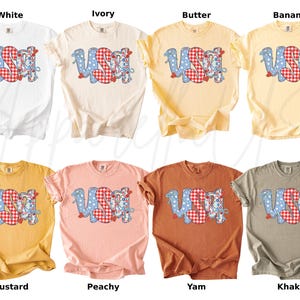 May include: A collection of eight t-shirts in various colors, including white, ivory, butter, banana, mustard, peachy, yam, and khaki. Each shirt features the word "USA" in a playful design with blue polka dots and red gingham patterns.