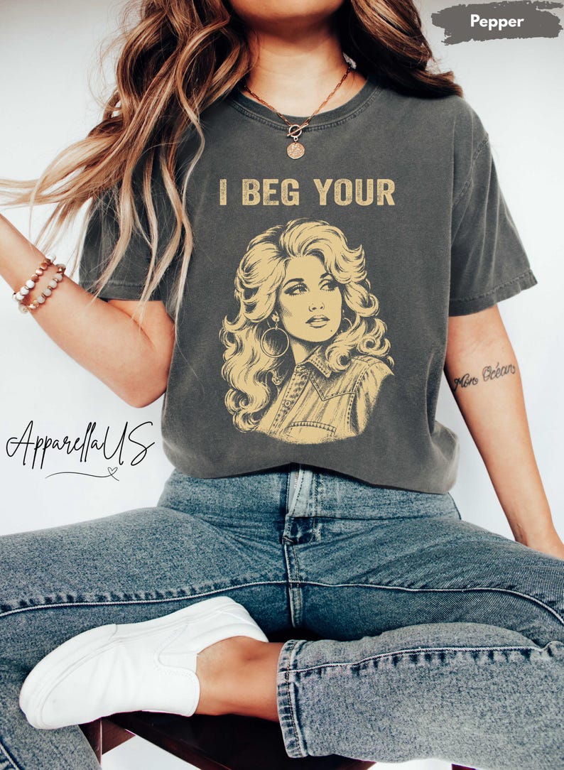 May include: A charcoal gray short-sleeve t-shirt featuring a yellow graphic illustration of a woman with voluminous hair and large hoop earrings. The text "I BEG YOUR" is printed in yellow above the illustration. The shirt has a crew neckline.