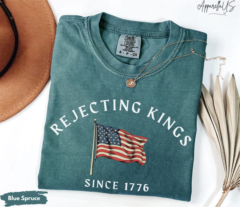 May include: Blue spruce t-shirt with the text "Rejecting Kings Since 1776" and an American flag graphic. The shirt is folded and has a Comfort Colors tag. A gold necklace with a pendant is on the shirt.