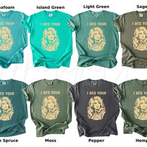 May include: A collection of eight short-sleeve t-shirts in various shades of green and gray, including seafoam, island green, light green, sage, blue spruce, moss, pepper, and hemp. Each shirt features a graphic of a woman and the text I BEG YOUR.