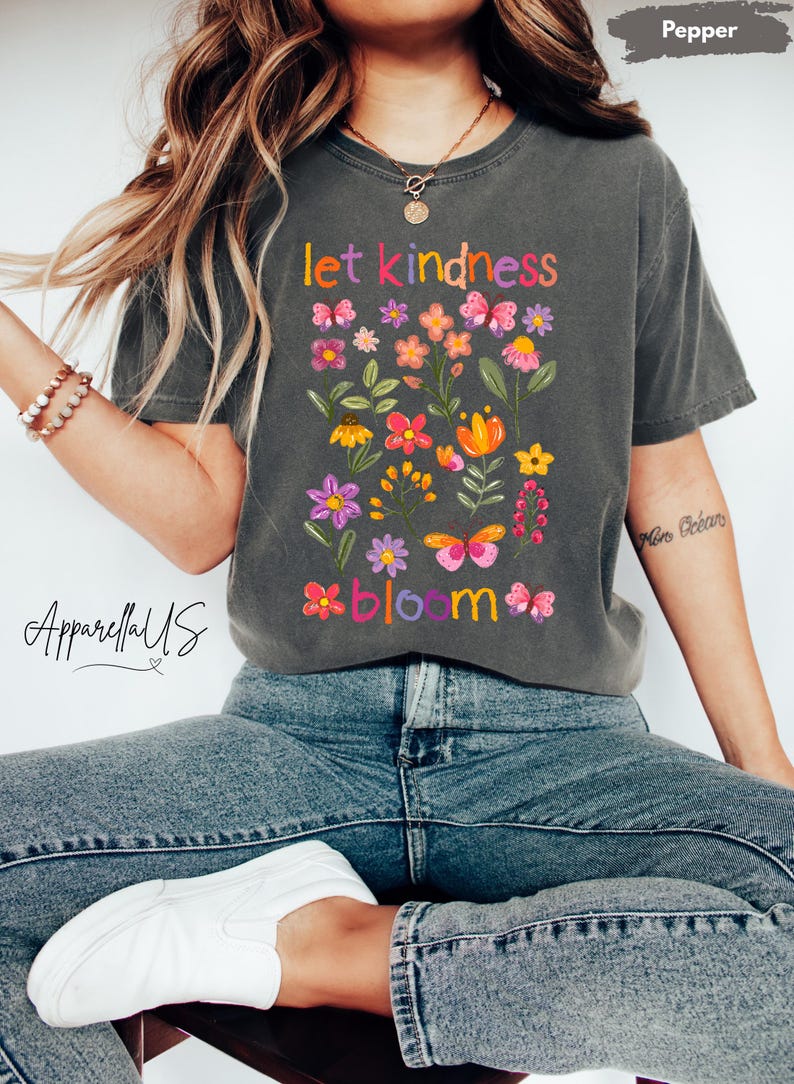 Pode incluir: T-shirt cinzenta escura com as palavras "let kindness bloom" em letras coloridas. A camisa apresenta um design floral com v&aacute;rias flores e borboletas. O nome da marca "ApparellUS" est&aacute; impresso no lado esquerdo.