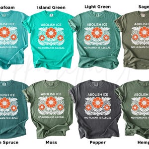 May include: A variety of t-shirts in different colors, including seafoam, island green, light green, sage, blue spruce, moss, pepper, and hemp. Each shirt features the text "ABOLISH ICE" above a floral design and the phrase "NO HUMAN IS ILLEGAL" below.