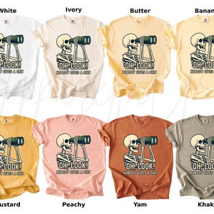 Op de afbeelding: Een collectie t-shirts in diverse kleuren, waaronder wit, ivoor en mosterd. Elk shirt heeft een afbeelding van een skelet met een verrekijker en de tekst "OH LOOK! NOBODY GIVES A SHIT."