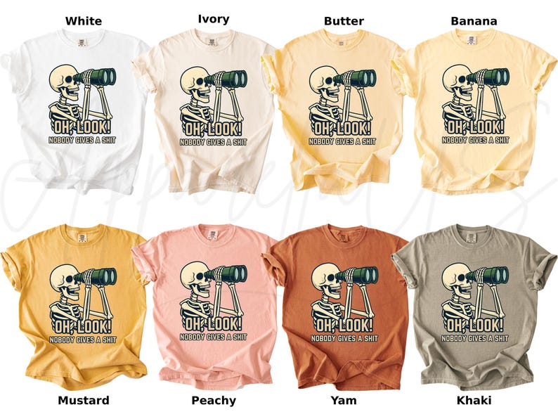 Op de afbeelding: Een collectie t-shirts in diverse kleuren, waaronder wit, ivoor en mosterd. Elk shirt heeft een afbeelding van een skelet met een verrekijker en de tekst "OH LOOK! NOBODY GIVES A SHIT."