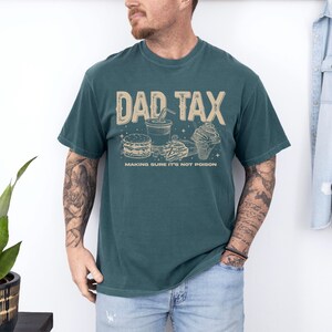 May include: Teal-colored t-shirt with the text "DAD TAX" in a tan font, along with illustrations of fast food items. The phrase "MAKING SURE IT'S NOT POISON" is printed below the graphic. The shirt is worn by a person.