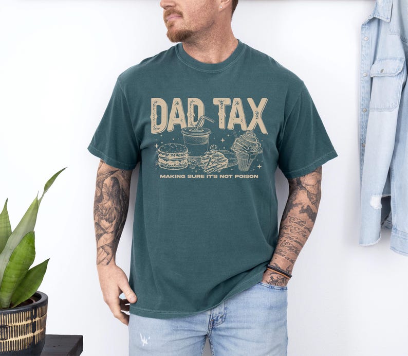 May include: Teal-colored t-shirt with the text "DAD TAX" in a tan font, along with illustrations of fast food items. The phrase "MAKING SURE IT'S NOT POISON" is printed below the graphic. The shirt is worn by a person.