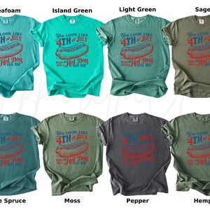 May include: Collection of t-shirts in various colors, each featuring a graphic with a hot dog and the text "You look like 4th of July makes me want a hot dog real bad". Colors include seafoam, island green, light green, sage, blue spruce, moss, pepper, and hemp.