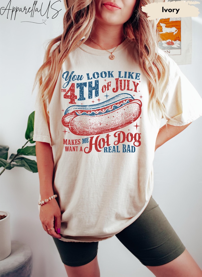 May include: Ivory-colored t-shirt with a patriotic hot dog graphic. The design features the text "You look like the 4th of July" and "Makes me want a Hot Dog real bad" in red and blue. A fun, casual tee for Independence Day.