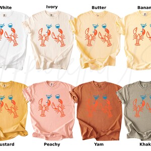 K&ouml;nnte beinhalten: T-Shirts in verschiedenen Farben, darunter Wei&szlig;, Elfenbein, Butter, Banane, Senf, Pfirsich, Yam und Khaki. Jedes Shirt zeigt eine Grafik von zwei Hummern, die Cocktails halten.